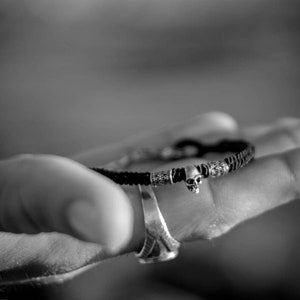 May include: Black and silver bracelet featuring a small skull charm. The bracelet has a woven black band with silver accents. The skull charm is centered on the bracelet. The bracelet is worn on a wrist.