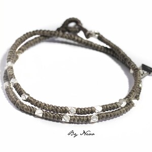 Könnte beinhalten: Ein doppeltes Armband aus braunem Kordel mit silbernen Perlen und einem silbernen Pfeilspitzenverschluss.