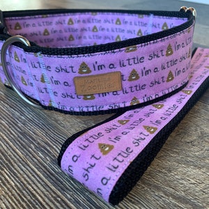 Puede incluir: Un collar para perro morado con ribete negro y una correa a juego. El collar y la correa están impresos con la frase "Soy una pequeña mierda" y un emoji de caca. El collar tiene un anillo en D de metal plateado.