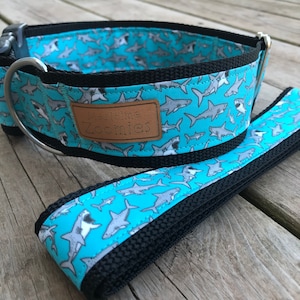 Pode incluir: Um conjunto de coleira e trela para cães azul e preto com um padrão de tubarão. A coleira tem um anel D de metal prateado e uma etiqueta de couro marrom que diz "Authentic Zoomies".