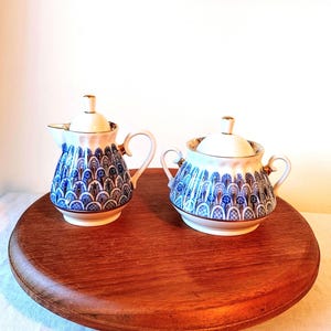 May include: A set of two porcelain creamer and sugar containers with blue and gold floral patterns. The containers are on a wooden surface.