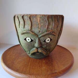 May include: A green ceramic face-shaped planter with white eyes and a moustache, sitting on a round wooden tray. The planter has a textured surface and a weathered appearance, suggesting an aged or antique aesthetic.