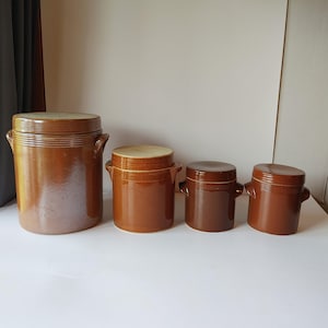 May include: A set of four graduated brown ceramic storage jars with lids. Each cylindrical jar features horizontal ribbed detailing near the top rim and two small side handles. The jars vary in size, ranging from a large vessel to three smaller ones.