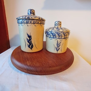 Peut inclure: Deux bocaux en céramique avec couvercles. Les bocaux sont de couleur crème avec des motifs floraux bleus et des motifs de feuilles. Ils sont exposés sur un support rond en bois. Les couvercles ont un bouton sur le dessus.