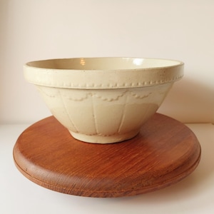 May include: A large, off-white ceramic mixing bowl with a scalloped design around the rim and a fluted pattern on the sides. The bowl is sitting on a wooden surface.