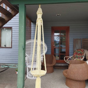 May include: A white macrame plant hanger with a tassel hanging from the bottom. The hanger is hanging from the ceiling of a porch with wicker chairs and a white wicker chair.