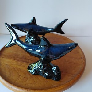 May include: Two ceramic shark figurines with a glossy, deep blue and black glaze. Each shark is mounted on a textured, dark, rock-like base, capturing the shark in a swimming position with fins extended and tails curved, showcasing a smooth finish.