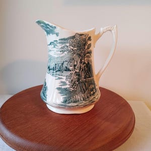May include: A white ceramic jug with a green and white landscape design. The jug has a handle and a spout.