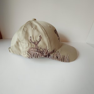 May include: Tan baseball cap with a brown embroidered deer and grass design. The cap has a curved brim and an adjustable strap. The cap is made of a durable material and is suitable for outdoor activities.