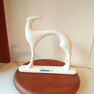 May include: A white ceramic sculpture of a stylized greyhound dog. The sculpture has a sleek, minimalist design with a curved body and long legs. The base has the text "Haeger Potteries USA".