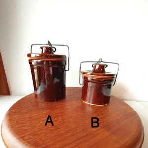Peut inclure: Deux pots en céramique marron avec couvercles et poignées en fil de fer. Les pots sont étiquetés A et B et sont posés sur une surface en bois. Les pots ont une finition brillante et un design classique.
