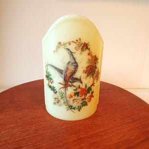 May include: A white glass candle holder with a floral design featuring a blue and brown bird with a long tail. The candle holder is shaped like a rounded rectangle.
