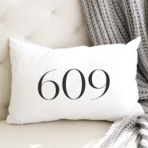 May include: White rectangular pillow with the number "609" printed in black. The pillow is on a light-colored, tufted surface, with a gray knitted blanket draped nearby. The pillow is a decorative home accent.