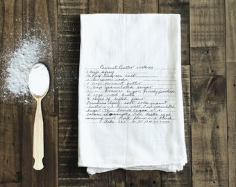 May include: A white kitchen towel with a handwritten peanut butter cookie recipe. The recipe includes ingredients like peanut butter, butter, sugar, eggs, and flour. The towel is on a wooden surface with a spoon and white powder.