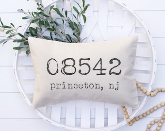 Può includere: Un cuscino rettangolare beige con la scritta "08542 Princeton, NJ" stampata in nero. Il cuscino è posto su un vassoio bianco intrecciato, con rami verdi e una ghirlanda di perline di legno.