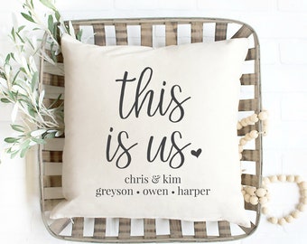 May include: A white throw pillow with black text that reads "This is us" with a small heart symbol. Below the text is "Chris & Kim Greyson Owen Harper". The pillow is in a woven basket with a wooden bead garland.