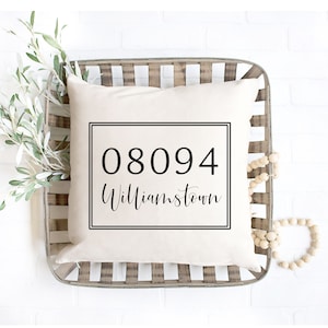 May include: A square, cream-colored pillow with the text "08094 Williamstown" in a black, framed design. The pillow is placed in a rustic, woven basket with white and gray stripes. Decorative greenery and wooden beads are arranged around the pillow.