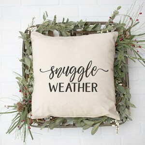 May include: A white throw pillow with black text that reads "snuggle weather" in a decorative font. The pillow is sitting in a woven basket with green leafy branches and red berries.