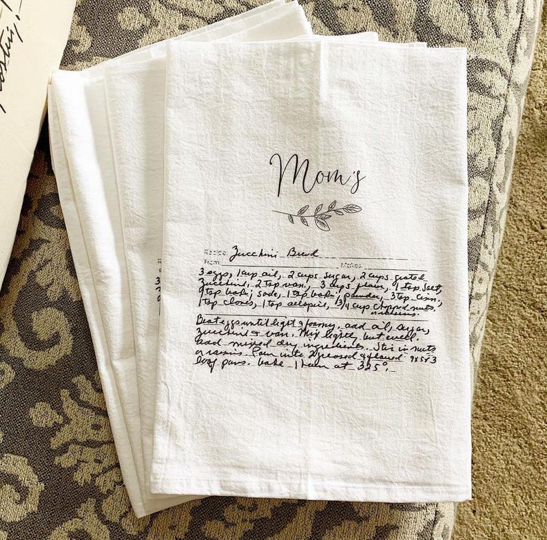 May include: White kitchen towel with a handwritten recipe for Zucchini Bread. The recipe is titled "Mom's" and includes ingredients like sugar, zucchini, vanilla, and spices. The towel is perfect for adding a personal touch to your kitchen.