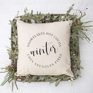 May include: A white throw pillow with black text that reads "Snowflakes Hot Cocoa Cozy Snuggles Fires" in a circle around the word "winter".