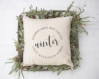 May include: A white throw pillow with black text that reads "Snowflakes Hot Cocoa Cozy Snuggles Fires" in a circle around the word "winter".
