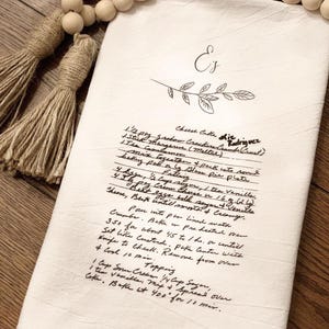 May include: A handwritten recipe for cheese cake on a white tea towel with the title "Cheese Cake die driguez". The recipe includes ingredients like graham cracker crust, butter, sugar, eggs, cream cheese, and vanilla. The recipe also includes instructions for baking and serving the cheese cake.