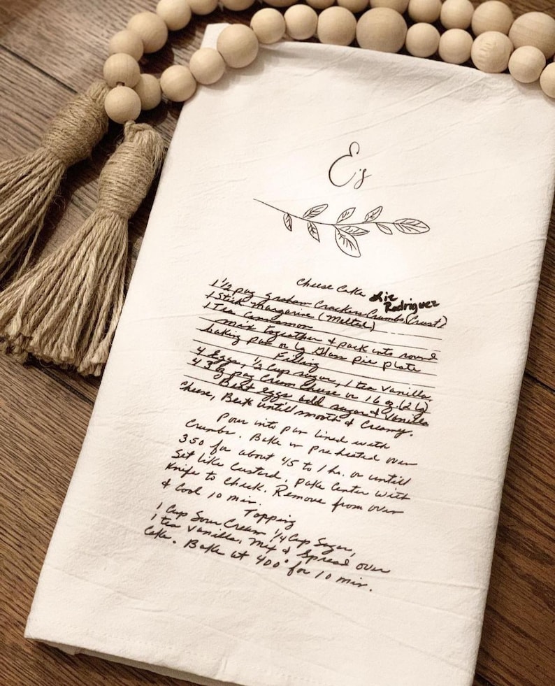 May include: A handwritten recipe for cheese cake on a white tea towel with the title "Cheese Cake die driguez". The recipe includes ingredients like graham cracker crust, butter, sugar, eggs, cream cheese, and vanilla. The recipe also includes instructions for baking and serving the cheese cake.
