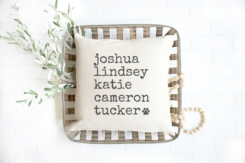 Pode incluir: Uma almofada branca com texto preto que diz "joshua lindsey katie cameron tucker" com um s&iacute;mbolo de pata. A almofada est&aacute; em uma cesta tecida com contas de madeira.