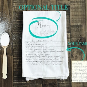 May include: A white tea towel with a handwritten peanut butter cookie recipe titled "Nana's". The recipe is written in black ink and includes ingredients like peanut butter, sugar, and eggs. The towel is on a wooden surface with a wooden spoon and a small pile of white powder.