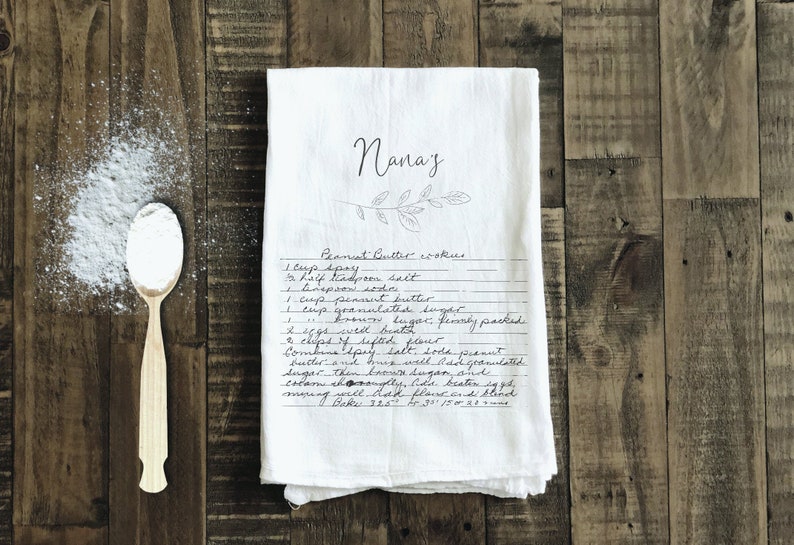 May include: A white tea towel with a handwritten recipe for peanut butter cookies. The recipe is titled "Nana's Peanut Butter Cookies" and includes ingredients like spry, salt, peanut butter, granulated sugar, flour, eggs, and baking soda.