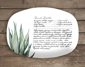 Può includere: Un piatto in ceramica bianca con una ricetta di peanut brittle scritta a mano. Il piatto presenta un'illustrazione ad acquerello di piante verdi.