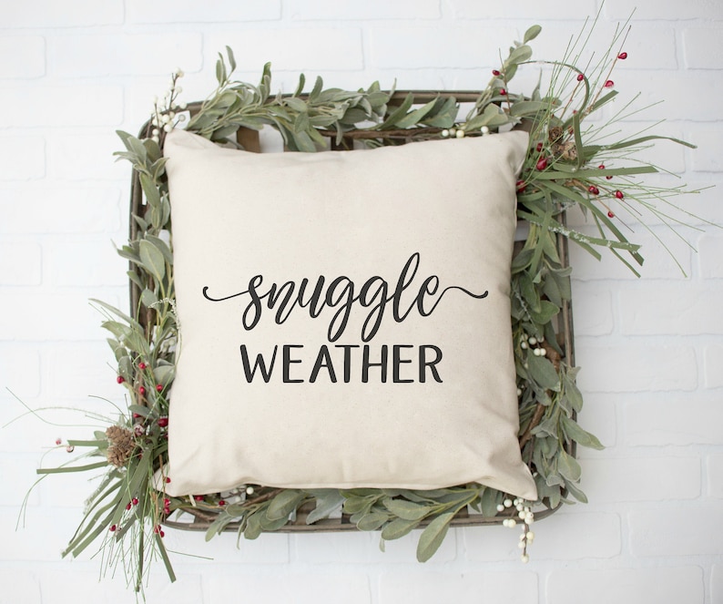 Pu&ograve; includere: Un cuscino bianco con testo nero che recita "snuggle weather" in un carattere decorativo. Il cuscino &egrave; seduto in un cesto intrecciato con rami verdi e bacche rosse.
