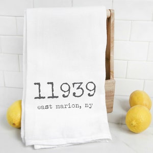 May include: White kitchen towel with black text that reads "11939 east marion, ny". The towel is hanging on a wooden rack and there are two yellow lemons in the foreground.