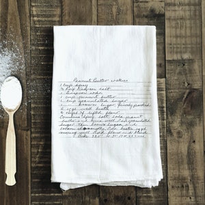 May include: A white tea towel with a handwritten peanut butter cookie recipe. The recipe includes ingredients like peanut butter, sugar, flour, and eggs. The towel is on a wooden surface with a spoon and a pile of white powder.
