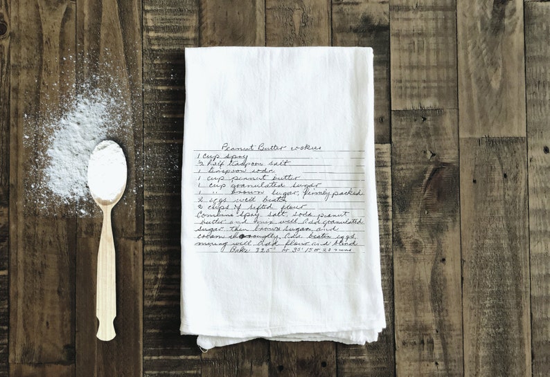 May include: A white tea towel with a handwritten peanut butter cookie recipe. The recipe includes ingredients like peanut butter, sugar, flour, and eggs. The towel is on a wooden surface with a spoon and a pile of white powder.