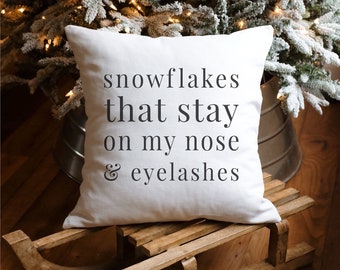 May include: A white square pillow with the text "snowflakes that stay on my nose & eyelashes" in black. The pillow is on a wooden sled, with a Christmas tree in the background.