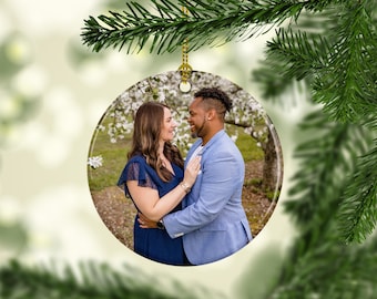 May include: A round, clear glass ornament with a photo of a couple in a garden setting. The couple is standing close together, smiling and looking at each other. The background is a blurred image of trees and flowers.