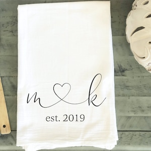May include: White kitchen towel with the initials "m" and "k" connected by a heart, and the year "est. 2019" printed in black. A wooden spoon and a decorative white pumpkin are also visible.