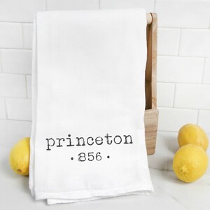 May include: White kitchen towel with black text that reads "princeton . 856 ." hanging on a wooden rack. Two yellow lemons are on a white countertop.