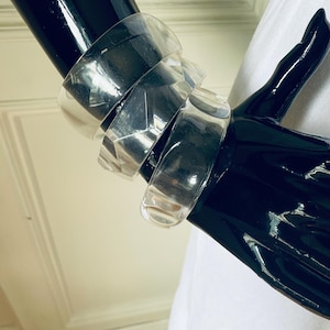 May include: A set of three clear, transparent acrylic bangle bracelets with a faceted, geometric design. The bangles are stacked together, showcasing a smooth, reflective surface and a rigid, circular structure that catches the light.