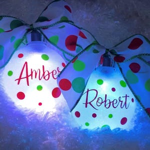 May include: Two illuminated Christmas ornaments with the names "Amber" and "Robert" in red script. The ornaments are decorated with red and green polka dots and topped with a white ribbon with matching polka dots. The background is a soft, glowing white.