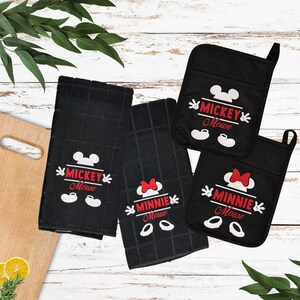 May include: Two black kitchen towels and two black pot holders featuring white and red graphics. One towel and one pot holder display the Mickey Mouse logo with ears and shoes. The other towel and pot holder display the Minnie Mouse logo with a bow.