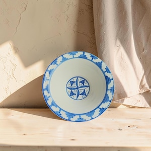 Op de afbeelding: Een ronde keramische kom met een witte basis en handgeschilderde blauwe details. De rand is versierd met een herhalend patroon van kleine blauwe voetstukvormen, terwijl het midden een cirkelvormig medaillon met een bloemmotief bevat.