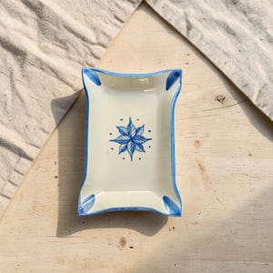 Op de afbeelding: Een rechthoekig keramieken dienblad met een crèmekleurige basis en gebogen randen. Het dienblad is voorzien van een handgeschilderd blauw bloemsterontwerp in het midden en blauwe accenten langs de vier hoeken voor een strakke uitstraling.