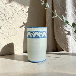 Può includere: Un bicchiere cilindrico in ceramica con una liscia smaltatura color bianco sporco. La parte superiore presenta una fascia decorativa blu dipinta a mano con un motivo ad archi ripetuti e piccoli punti. La base è rifinita da una sottile linea blu.