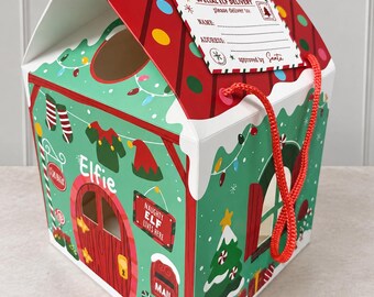 May include: A house-shaped cardboard gift box with a red roof and green walls featuring Christmas illustrations. The box includes a tag labeled SPECIAL ELF DELIVERY, a red rope handle, and printed details like a door, mail slot, and tree.