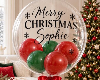 May include: A large, clear, spherical balloon filled with smaller red and green balloons. The exterior is printed with the black text Merry CHRISTMAS Sophie and black snowflake icons. A bright red satin ribbon is tied at the base.
