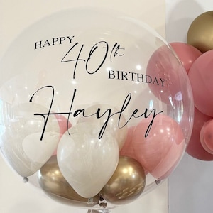 May include: A clear bubble balloon with the text "Happy 40th Birthday Hayley" and smaller balloons inside. The smaller balloons are white, gold, and pink.
