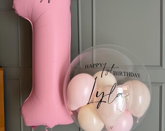 May include: A large matte pink number one balloon personalized with the name Lyla in black script, paired with a clear round balloon filled with smaller pink and cream balloons, featuring the black text Happy 1st Birthday and the name Lyla.
