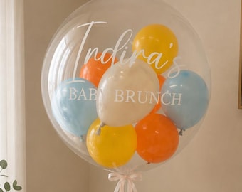 May include: A large clear balloon filled with smaller balloons in yellow, orange, white, and blue. The balloon is printed with the text "Indira's BABY BRUNCH" in a delicate script. Long, pale pink ribbons are attached to the base of the balloon.